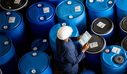 Ein Mitarbeiter von REMONDIS mit Helm steht inmitten vieler blauer Fässer und hält ein Tablet in der Hand ALT: Ein Person mit Haube, Schutzbrille, einfachem Mundschutz und Nitrilhandschuhen entnimmt mit einer Spritze eine Flüssigkeit aus einer kleinen Durchstechflasche. inmitten vieler blauer Fässer und hält ein Tablet in der Hand.