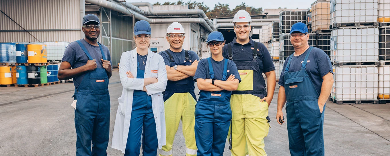 Eine Gruppe von REMONDIS-Mitarbeiten – 2 Frauen und 4 Männer – stehen in Arbeits- und Sicherheitskleidung auf einem Wertstoffhof nebeneinander und schauen in die Kamera.