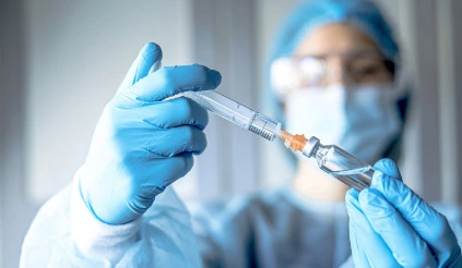 Ein Person mit Haube, Schutzbrille, einfachem Mundschutz und Nitrilhandschuhen entnimmt mit einer Spritze eine Flüssigkeit aus einer kleinen Durchstechflasche.